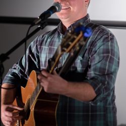 Eli Conley - Image Description: Eli, a white man, is smiling on stage at a microphone with a guitar.