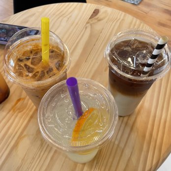 Soule' Coffee + Bubble tea - Three iced beverages with clear plastic lids and colorful straws (yellow, purple, black and white) are arranged on a round wooden table. The drinks include a coffee or tea with ice, a lemon-flavored drink with a purple straw, and a layered coffee drink with a black and white striped straw. The background shows part of the cafe interior with chairs and a patterned rug. - Coffee & Tea Near Me - Jackson, Mississippi