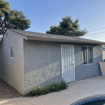 Acosta Plastering - Pool house.  The detail around the doors and windows was outstanding. - A gray stucco pool house with a metal security door and a window, surrounded by potted plants and a concrete patio. - Stucco Services Near Me - Gilbert, Arizona