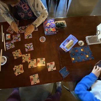 Maru Plus - Two children are engaged in a card game at a wooden table, with game cards, a Pictionary box, and other game accessories visible. - Japanese Near Me - Tucson, Arizona