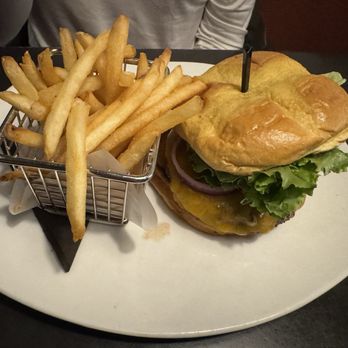 110 Grill - Cheeseburger and fries - A cheeseburger with a bun, lettuce, tomato, onion, and melted cheese is presented on a white plate. A small metal basket filled with French fries accompanies the burger. The plate is placed on a dark table, and part of a person wearing a light-colored shirt is visible in the background. - American Near Me - Hadley, Massachusetts