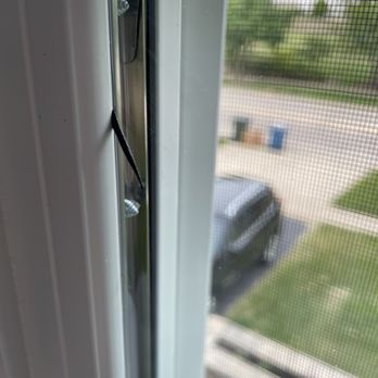 Friendly Windows - Rubbery material coming out of a window - The image depicts a close-up of a window screen with a visible tear or hole in the mesh material. The screen is part of a window frame, and through the mesh, an outdoor scene with a parked vehicle, some greenery, and a few objects on the ground can be seen. The focus is on the damaged screen, with the outdoor background slightly blurred. - Windows Installation Near Me - Barrington, Illinois