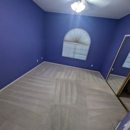 The image shows an empty bedroom with purple painted walls, a ceiling fan with attached lights, and a window with blinds that are partially open. The floor is carpeted with a light-colored carpet, and there is a mirrored closet door on one side of the room. The room appears clean and well-maintained, with electrical outlets visible on the walls.