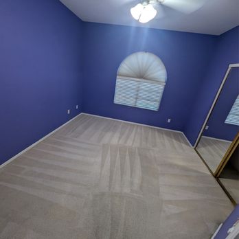 Steam Master Cleaning - The image shows an empty bedroom with purple painted walls, a ceiling fan with attached lights, and a window with blinds that are partially open. The floor is carpeted with a light-colored carpet, and there is a mirrored closet door on one side of the room. The room appears clean and well-maintained, with electrical outlets visible on the walls. - Carpet Cleaning Near Me - Phoenix, Arizona