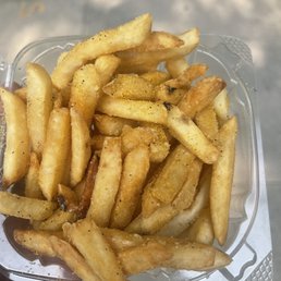 Classic Luv Fries - A transparent plastic container filled with French fries seasoned with salt and black pepper. The fries are cut in a standard shape, with some showing a golden-brown color and crispy texture. The container is held by a hand, and the background is blurred, indicating an outdoor setting.
