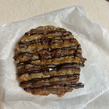 Cookie Time Bakery - Toffee Turtle (yum!!) - A cookie with a rough, irregular shape is decorated with chocolate and caramel drizzles, placed on parchment paper. - Bakeries Near Me - Arlington, Massachusetts