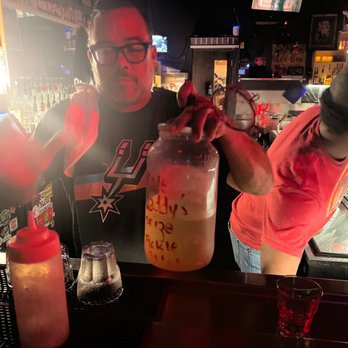 The Side Bar - The dream team - A man with glasses and dark hair is standing behind a bar counter, holding a large jar with a drink and a small toy lobster on top. The bar area has bottles of alcohol, bar tools, and a red squeeze bottle. The background shows a dimly lit bar with a television screen and various decorations. The man appears to be preparing or presenting a drink. - Bars Near Me - Austin, Texas