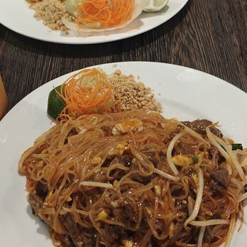 Sukhothai - Beef pad thai - Two plates of Pad Thai are on a dark wooden table, each with different ingredients, garnished with lime, shredded carrots, and crushed peanuts. - Thai Near Me - Albany, New York