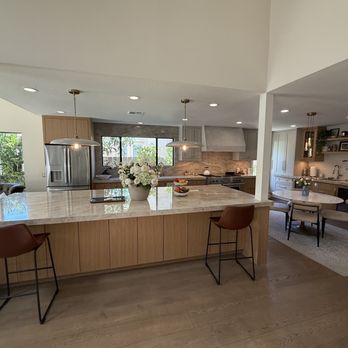PCD Remodeling - The image shows a contemporary kitchen with a large marble countertop island, pendant lighting, and a dining area with a round table and chairs. The kitchen includes a stainless steel refrigerator, a stove with a range hood, and open shelving with decorative items. Large windows provide natural light, and the space has a neutral color palette with wood accents. - General Contractors Near Me - La Jolla, California