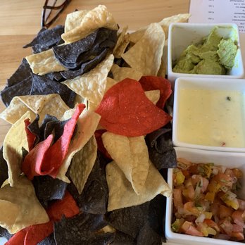 The View Pub - Chips and dip - A plate of multicolored tortilla chips served with guacamole, queso, and pico de gallo salsa on a wooden table, with a pair of glasses nearby. - New American Near Me - Burke, Vermont