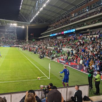 Q2 Stadium - A soccer match is taking place at Q2 Stadium, with a player in a blue jersey and the number 7 standing near the corner flag on the field. The stadium is filled with spectators seated in multiple tiers, and bright stadium lights illuminate the scene. Photographers and camera operators are positioned along the sideline capturing the game. - Stadiums & Arenas Near Me - Austin, Texas