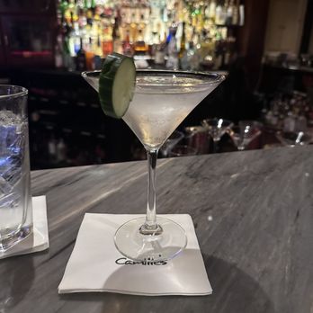 Camille's - A clear martini with a lime wedge garnish is positioned on a white napkin on a gray marble bar counter. The background features a blurred bar area with various bottles and bar equipment, indicating an upscale Italian restaurant setting at Camille's in Providence, RI. The image captures the drink's presentation and the bar's ambiance. - Italian Near Me - Providence, Rhode Island