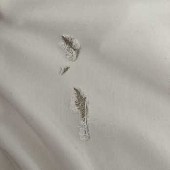 The Laundry Wheel - Holes in sheet - The image depicts a piece of white fabric with three visible tears, each with frayed edges and loose threads. The fabric appears to be woven, with a smooth surface, and the tears are irregular in shape and size. The background is plain, emphasizing the damaged areas of the fabric. - Laundromat Near Me - Phoenix, Arizona