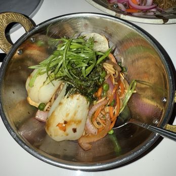 Sanctuary - Chow mein - A metal bowl holds chow mein with vegetables such as peas, carrots, onions, and greens, along with noodles. The dish is garnished with fresh greens on top. The bowl is on a white surface, with other dishes partially visible in the background. - Cocktail Bars Near Me - Chicago, Illinois