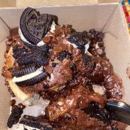 Chocolate Overload - A dessert with a mixture of chocolate chips, Oreo cookies, and brownie pieces served in a small cardboard box. The Oreo cookies are broken into pieces and placed on top, along with chocolate chips. There are also chunks of brownie with a chocolate topping. A spoon is partially visible in the lower right corner, indicating the dessert is ready to be eaten.