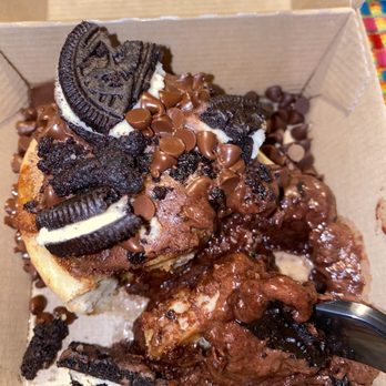 Cinnaholic - Chocolate Overload - A dessert with a mixture of chocolate chips, Oreo cookies, and brownie pieces served in a small cardboard box. The Oreo cookies are broken into pieces and placed on top, along with chocolate chips. There are also chunks of brownie with a chocolate topping. A spoon is partially visible in the lower right corner, indicating the dessert is ready to be eaten. - Bakeries Near Me - Gambrills, Maryland
