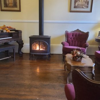 Murphys Historic Hotel - Lobby - A living room features vintage-style purple upholstered chairs and a sofa, a wooden coffee table with a floral arrangement, a wood stove with a fire, and decorative lamps and pictures. - Hotels Near Me - Murphys, California