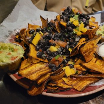 Cafecito - A plate of nachos features crispy, golden-brown tortilla chips topped with black beans, melted cheese, and diced vegetables. The dish is served with sides of guacamole and sour cream, placed in small plastic containers. The presentation is on a round ceramic plate with a paper liner, set on a dark wooden table. - Cuban Near Me - Chicago, Illinois