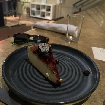 Sava's - A slice of cheesecake topped with berries and an edible flower is placed on a black plate on a restaurant table, with sunglasses, a glass, and a napkin nearby. - Bars Near Me - Ann Arbor, Michigan