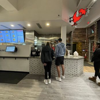 CHiKN - Two customers, a woman and a man, stand at a counter in a fast-food restaurant, placing their orders. The counter has a patterned front, and digital menu screens display food options above. A staff member is behind the counter assisting. The restaurant interior features a modern design with wood and metal accents, and a rooster logo is mounted on the ceiling. Other customers are visible in the background, and the floor is covered with wood-look flooring. - Chicken Shop Near Me - Crafton, Pennsylvania