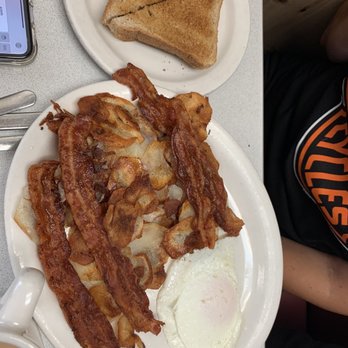 Bud Willman's - Bacon, eggs, American fries and toast - A breakfast meal featuring several strips of crispy bacon, a fried egg with a runny yolk, and a serving of potato chips on a white plate. There is also a side of toasted bread cut into triangles on a separate plate, and a cup of coffee partially visible in the corner. The setting appears to be a casual diner or restaurant. - Burgers Near Me - Manitowoc, Wisconsin