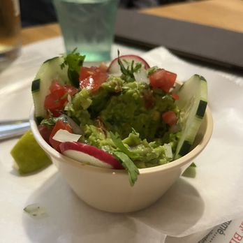 Los Arcos - Guacamole - A small bowl of guacamole topped with diced tomatoes, radishes, and cucumber slices, with a lime wedge on the side, on a table at a restaurant. - Tacos Near Me - Chicago, Illinois