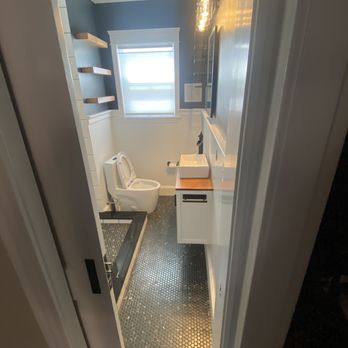 Victor's Finish Work - To show size of bathroom - A compact bathroom with dark hexagonal floor tiles, a white toilet, a white vanity with a vessel sink, and wall-mounted shelves. A window provides natural light. - Refinishing Services Near Me