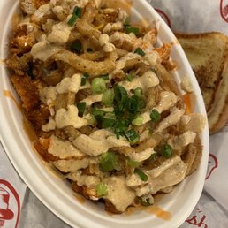 Grilled Buffalo Chicken Wrap - A food dish featuring Buffalo Chicken Mac and Cheese in a white oval bowl. The dish is topped with chopped green onions, crispy fried onion strings, and drizzled with a creamy sauce. In the background, there are slices of toasted bread on branded paper with red logos and text. The presentation suggests a comfort food item served at a casual dining or fast-food restaurant.