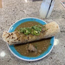 Their tasty burrito as is - A burrito with a toasted, slightly charred tortilla is served on a blue-rimmed plate, topped with microgreens and sliced jalapeños, sitting in a green sauce. A mason jar containing a light-colored drink with a straw is positioned behind the plate. The setting appears to be a casual dining environment with a granite or stone countertop.