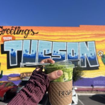 Brucha - Banana Cream Matcha - A person wearing a purple long-sleeve garment holds a plastic cup of green matcha drink with a straw, in front of a colorful mural on a brick wall in Tucson, Arizona. The mural features large, stylized text and bright colors, with a parking lot and cars visible at the bottom of the image. The sky is clear and blue. - Gelato Near Me - Tucson, Arizona