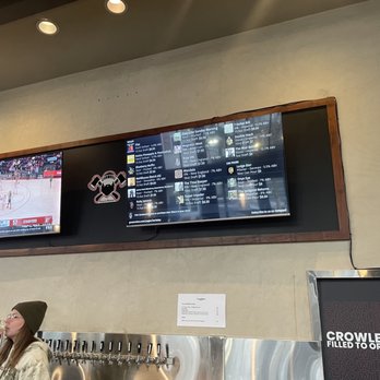 Great Notion - Berkeley - Two digital screens are mounted on a wall inside a brewery or bar. The left screen displays a basketball game, while the right screen shows a beer menu with various craft beers and their details. A woman wearing a beanie and glasses is drinking from a glass in the foreground, standing near a beer tap system with multiple taps. The wall has a neutral color, and there is a small sign below the screens and a black sign with the word 'CROWLERS' on the right side. - Brewpubs Near Me - Berkeley, California