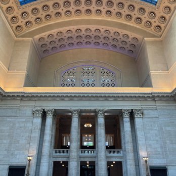 Amtrak - Chicago Union Station - The image shows the interior of a large, historic building with a high ceiling and classical architectural elements. Tall, ornate columns support the upper level, and decorative ceiling details are visible. A large glass skylight at the top allows natural light to illuminate the space. The floor is polished, and there are vintage-style lamps along the walls. The overall design emphasizes grandeur and elegance. - Trains Near Me - Chicago, Illinois