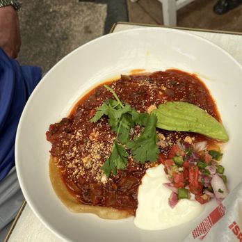 Centro Mexican Kitchen - Huevos Rancheros - The image shows a plate of huevos rancheros served with eggs covered in red salsa, garnished with cilantro, a slice of avocado, and a dollop of sour cream. The dish is presented on a white plate on a table, with some restaurant furniture visible in the background. - Mexican Near Me - Boulder, Colorado