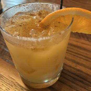 Bob's Seafood - Painkiller - A beverage in a clear glass contains a yellow-orange cocktail with ice cubes and a slice of orange garnishing the rim. The drink appears frothy on top, with a straw inserted. The glass is placed on a wooden table, and the background is slightly blurred, indicating an indoor setting. - Seafood Markets Near Me - Windham, Maine