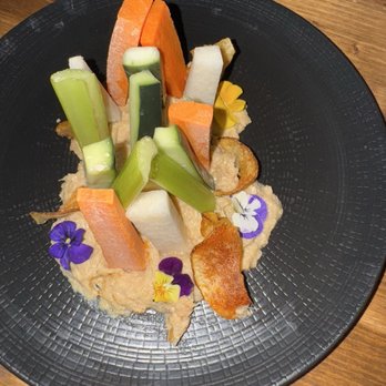 3128 Social House - Double duper - A presentation of assorted vegetable sticks including cucumber, carrot, and jicama, arranged on a black plate. The dish is garnished with potato chips and edible flowers, served on a wooden table. The plate has a textured surface, and a wine glass is partially visible in the background. - Gastropubs Near Me - Albuquerque, New Mexico