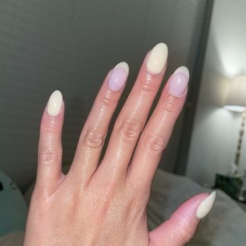 Nails by District - Yellow dip with Thao - A close-up of a hand with manicured nails, featuring a combination of pastel colors such as cream and light purple. The nails are shaped in an almond style, and some have a subtle design or gradient effect. The background shows an indoor environment with a gray wall and a lamp, indicating a setting like a nail salon or a home. The skin appears moisturized and well-maintained. - Nail Salons Near Me - Washington, DC, District of Columbia