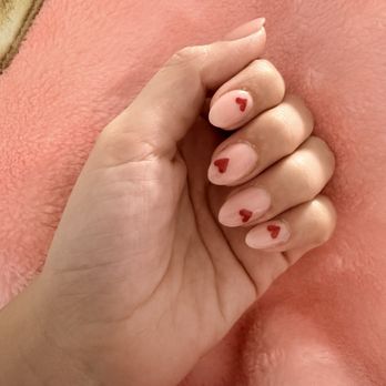 Sparkle Nails - Valentines set - The image shows a close-up of a hand with five fingers, each nail painted with a clear or light base coat and decorated with small red heart designs. The background appears to be a soft, pinkish textured surface, possibly a blanket or fabric. The focus is on the nails and the hand's natural skin tone, with no additional objects or context visible. - Nail Salons Near Me - Ceres, California