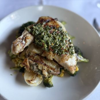 Haven Restaurant and Bar - Spice Roasted Springer Mountain Chicken - A roasted chicken leg with crispy skin is topped with a green herb sauce, possibly chimichurri or pesto. The dish is served on a white plate with vegetables and grains underneath, including broccoli, corn, and possibly rice or quinoa. The presentation emphasizes the roasted chicken and fresh herb topping, with a focus on the food's textures and colors. - Southern Near Me - Atlanta, Georgia