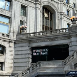 The image shows a historic, ornate building with detailed architectural features, including a large arched window with a clock. A sign for Wendella Boat Rides is visible above a staircase leading to an entrance. Several people are gathered at the bottom of the stairs, some wearing hats and casual clothing. The building has decorative elements, stone balustrades, and street lamps, indicating a prominent location in Chicago.