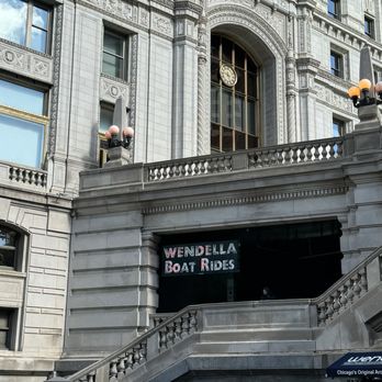 Chicago Architecture Center - The image shows a historic, ornate building with detailed architectural features, including a large arched window with a clock. A sign for Wendella Boat Rides is visible above a staircase leading to an entrance. Several people are gathered at the bottom of the stairs, some wearing hats and casual clothing. The building has decorative elements, stone balustrades, and street lamps, indicating a prominent location in Chicago. - Historical Tours Near Me - Chicago, Illinois