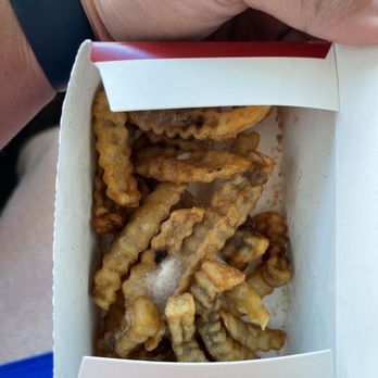 Zaxbys - Molded fries from 2nd kids meal. - A person is holding a white and red cardboard box filled with crinkle-cut French fries. Several fries inside the box show signs of mold, with fuzzy white patches. The person's hand, wearing a dark wristband, is visible gripping the box, and part of their arm and clothing are also seen. The background is blurred, focusing on the fries and the mold issue. - Fast Food Near Me - Columbus, Mississippi