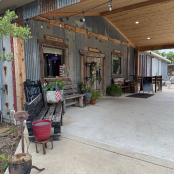 Oscar Store - Oscar store entry - The photo depicts the front exterior of a rustic dining venue built with corrugated metal and wood siding, featuring outdoor seating areas with benches and tables. Decorative signs with menu items are mounted above the windows, and string lights hang across the porch. Potted plants and vintage-style furniture add to the country aesthetic. The space includes a covered porch with wooden ceiling panels and outdoor lighting, creating a welcoming atmosphere. - American Near Me - Temple, Texas