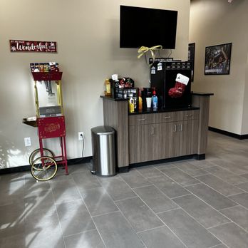 Caliber Collision - Complimentary coffee and tea service as well as fresh popcorn - The image shows a refreshment station in a waiting area, with a popcorn machine on a red cart, a countertop with drinks and condiments, and holiday decorations such as a stocking hanging from the ceiling and ornaments. A flat-screen TV is mounted on the wall above the station, and framed pictures are visible on the walls. The floor is tiled, and a trash can is placed next to the counter. - Body Shops Near Me - Rio Rancho, New Mexico