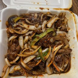 Mongolian Beef - A foam takeout container displays Mongolian beef with sliced onions, green peppers, and carrots coated in a dark, savory sauce. The food appears partially eaten, with some sauce and vegetable pieces visible around the edges of the container.