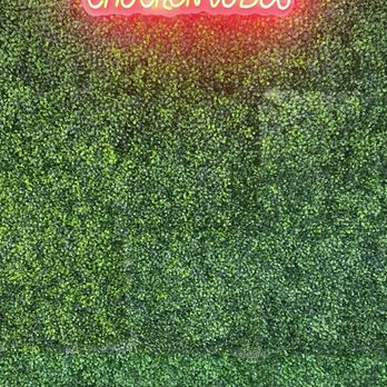 Midwestwings - Indoor decor - A bright pink neon sign displaying 'CHICKEN WINGS' is mounted on a lush green artificial hedge wall, with a black and white checkered floor at the bottom. - Chicken Wings Near Me - Phoenix, Arizona