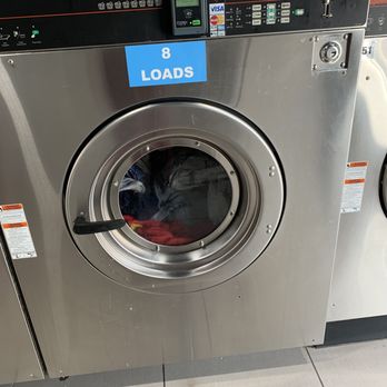 Spin Cycle Laundry Lounge - A stainless steel front-loading washing machine in a laundromat, featuring a digital control panel, a coin slot, and a glass door through which laundry inside is visible. The machine has a blue sticker indicating it can handle "8 LOADS" of laundry. The control panel displays a cycle time of 18 minutes and various payment options, including card readers. The floor is tiled, and part of another washing machine is visible beside it. - Laundromat Near Me - Manteca, California
