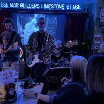 The Quarry Thornton - A band is performing live on stage at Del Mar Builders Limestone Stage, with three musicians visible: two guitarists and a drummer. The stage has a neon sign overhead, and a screen behind the band shows a person in a white outfit. Audience members are seated at tables in front of the stage, watching the performance. The setting appears to be an indoor venue with posters and decorations on the walls. - Bars Near Me - Thornton, Illinois