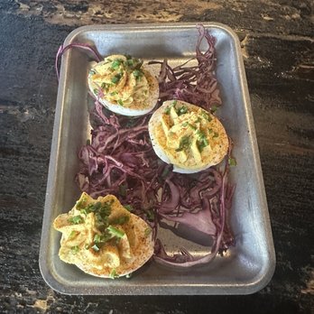 Dinosaur Bar-B-Que - Three deviled eggs topped with chopped green onions are placed on a rectangular metal tray. The tray also contains purple shredded cabbage as a garnish or decoration. The tray is set on a dark, textured wooden surface, and the eggs are evenly spaced with a simple presentation. - Barbeque Near Me - Troy, New York