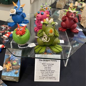 Ursel's Web - Multiple handcrafted wooden dragon figurines in colors such as blue, pink, red, green, and white are displayed on a glass table at an art or craft show. Each dragon has distinct features and expressions, with some holding accessories or decorated with small details. A printed label identifies the collection as 'The Dragon Family' with names and descriptions for each figure. Additional items and displays are visible in the background, indicating an artisan market setting. - Art Galleries Near Me - Middletown, Connecticut