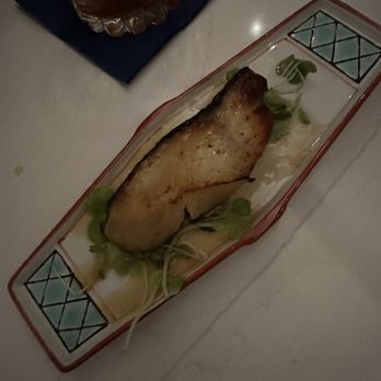 C Suite - Miso cod - A piece of grilled fish with a golden-brown exterior is placed on a rectangular ceramic plate featuring a geometric pattern in the corners. The fish is garnished with fresh greens and placed on a bed of sauce or dressing. In the background, there is a glass of beverage on a dark napkin, set on a white table surface. The lighting is dim, suggesting a restaurant setting. - Cocktail Bars Near Me - Toronto, Ontario